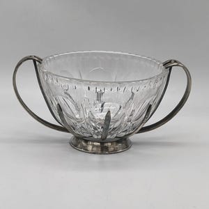 May include: A clear, cut-glass bowl with a silver-coloured metal base and handles. The bowl features a detailed pattern of vertical lines and floral designs. The handles curve outward from the bowl's rim, adding a decorative touch to this elegant piece.