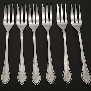 May include: Six silver forks with tines, long handles, and decorative detailing. Each fork has a floral design at the base of the handle. The forks are arranged on a black surface.