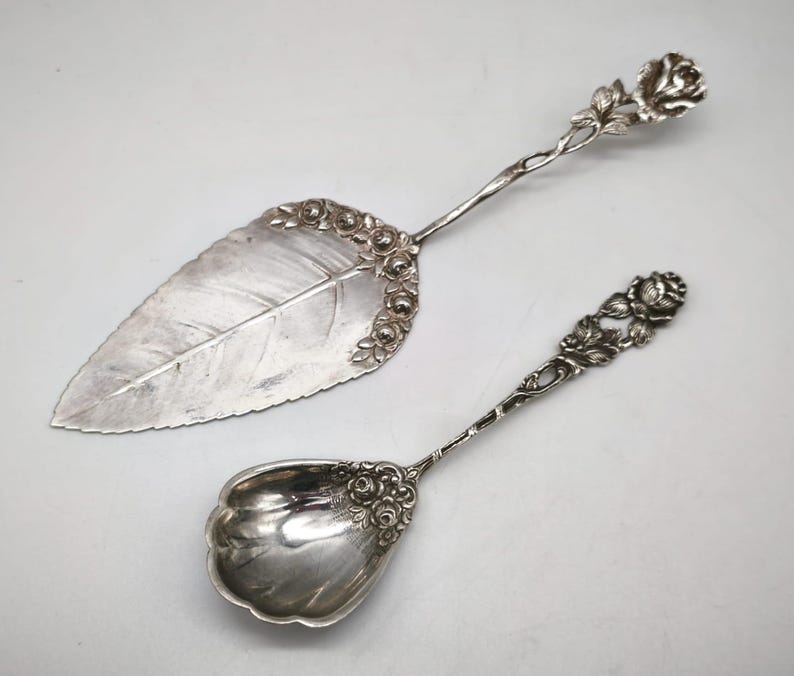 Peut inclure: Ensemble de service argent&eacute; comprenant une pelle &agrave; g&acirc;teau et une cuill&egrave;re. La pelle &agrave; g&acirc;teau a une lame en forme de feuille avec des d&eacute;tails de roses, tandis que la cuill&egrave;re a un bol arrondi et des accents de roses. Les deux pi&egrave;ces ont des manches orn&eacute;s.