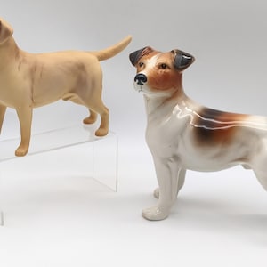 Puede incluir: Dos figuras de perros de cerámica. Uno es un Labrador Retriever marrón claro, y el otro es un Jack Russell Terrier con pelaje blanco, marrón y negro. El Labrador está sobre un expositor de acrílico transparente.