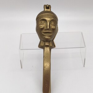 May include: A vintage brass nutcracker with a face design. The nutcracker features a detailed face with a smiling expression, a decorative collar, and a long handle. The brass has a weathered appearance.