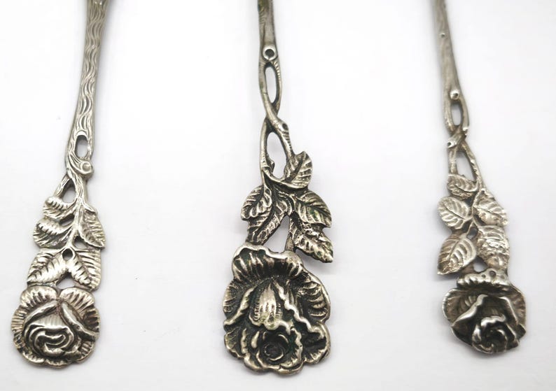 May include: Three antique silver spoons with intricate floral rose and leaf designs. The spoons have detailed stems and rose-shaped bowls, showcasing a vintage aesthetic.