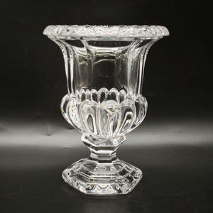 May include: A clear, ornate glass vase with a wide, scalloped rim and a pedestal base. The vase features a detailed, textured design and is likely made of crystal. The overall shape is reminiscent of a chalice or goblet.