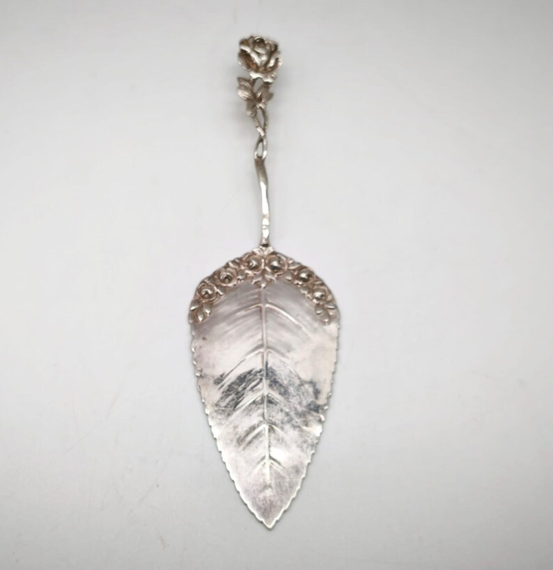 Peut inclure: Une pelle &agrave; g&acirc;teau argent&eacute;e avec une lame en forme de feuille et une poign&eacute;e en forme de rose. La lame pr&eacute;sente un motif de nervures de feuilles d&eacute;taill&eacute; et est bord&eacute;e de petits accents floraux. La poign&eacute;e est fine et torsad&eacute;e.