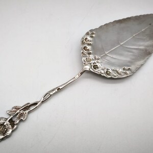 Peut inclure: Une pelle &agrave; g&acirc;teau argent&eacute;e avec une lame en forme de feuille et un motif floral sur le bord. La poign&eacute;e torsad&eacute;e se termine par une fleur, cr&eacute;ant une esth&eacute;tique &eacute;l&eacute;gante et vintage.