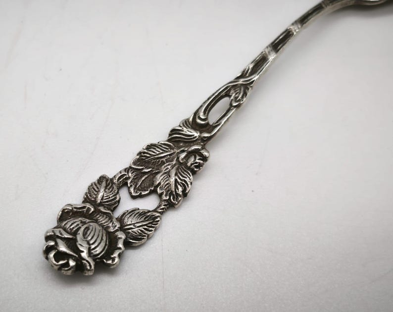 Peut inclure: Gros plan d'une cuill&egrave;re en argent avec un motif de rose. Le manche de la cuill&egrave;re pr&eacute;sente un motif complexe de feuilles et une rose d&eacute;taill&eacute;e &agrave; la base. La cuill&egrave;re est en m&eacute;tal brillant.