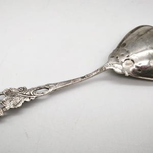 Peut inclure: Une cuill&egrave;re de service en argent avec un motif floral sur le manche. La cuill&egrave;re a un grand bol arrondi et une poign&eacute;e orn&eacute;e avec une rose et des feuilles. La cuill&egrave;re est probablement vintage.
