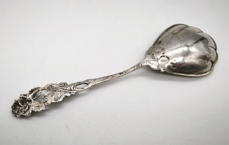Peut inclure: Une cuill&egrave;re de service en argent avec un motif floral sur le manche. La cuill&egrave;re a un grand bol arrondi et une poign&eacute;e orn&eacute;e avec une rose et des feuilles. La cuill&egrave;re est probablement vintage.