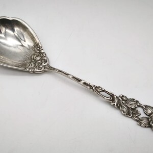 Peut inclure: Une cuill&egrave;re de service en argent avec un motif floral. La cuill&egrave;re a un grand bol festonn&eacute; et un long manche avec des d&eacute;tails de roses et de feuilles complexes. La cuill&egrave;re est probablement en argent sterling.