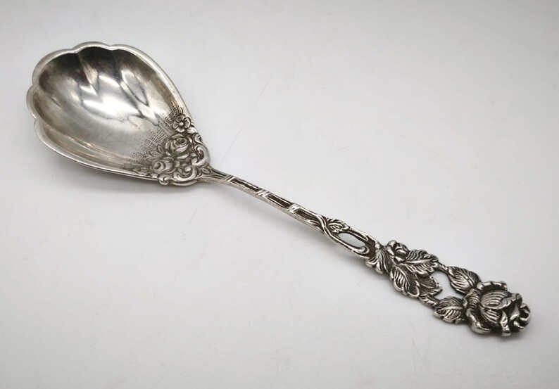 Peut inclure: Une cuill&egrave;re de service en argent avec un motif floral. La cuill&egrave;re a un grand bol festonn&eacute; et un long manche avec des d&eacute;tails de roses et de feuilles complexes. La cuill&egrave;re est probablement en argent sterling.