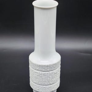 Könnte beinhalten: Weiße Keramikvase mit einem strukturierten Design um die Mitte. Die Vase ist hoch und schlank.