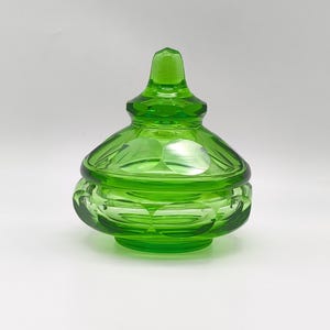 Puede incluir: Un tarro de vidrio verde brillante con tapa. El tarro tiene una base redonda y un patrón decorativo de círculos. La tapa es cónica con un pequeño pomo. El vidrio tiene un acabado brillante.