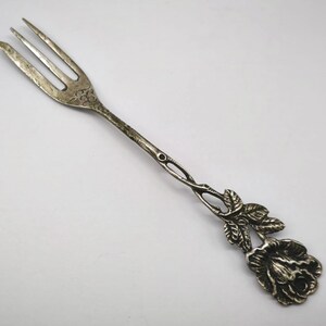 May include: Silver-toned fork with three tines. The handle features a decorative rose and leaf design. The fork is likely vintage, with a detailed floral motif, and is suitable for serving appetizers or desserts.