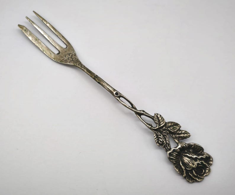 May include: Silver-toned fork with three tines. The handle features a decorative rose and leaf design. The fork is likely vintage, with a detailed floral motif, and is suitable for serving appetizers or desserts.