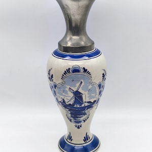 May include: A tall, decorative vase with a white ceramic body and a silver-colored metal top. The vase features a blue and white windmill scene, with blue floral accents. The base and rim have blue bands.