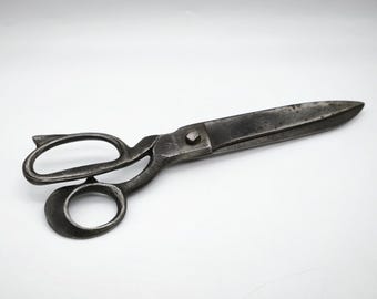 May include: A pair of antique metal scissors with a dark, aged appearance. The scissors have large finger holes and a long, pointed blade. The metal has a worn, textured look, suggesting age and use. The scissors are set against a plain white background.