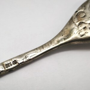 May include: Close-up of a silver serving utensil, possibly a fork or spoon, with ornate scrollwork and a hallmark stamp. The utensil has a tarnished appearance, suggesting age. The hallmark reads "835" and "CP".