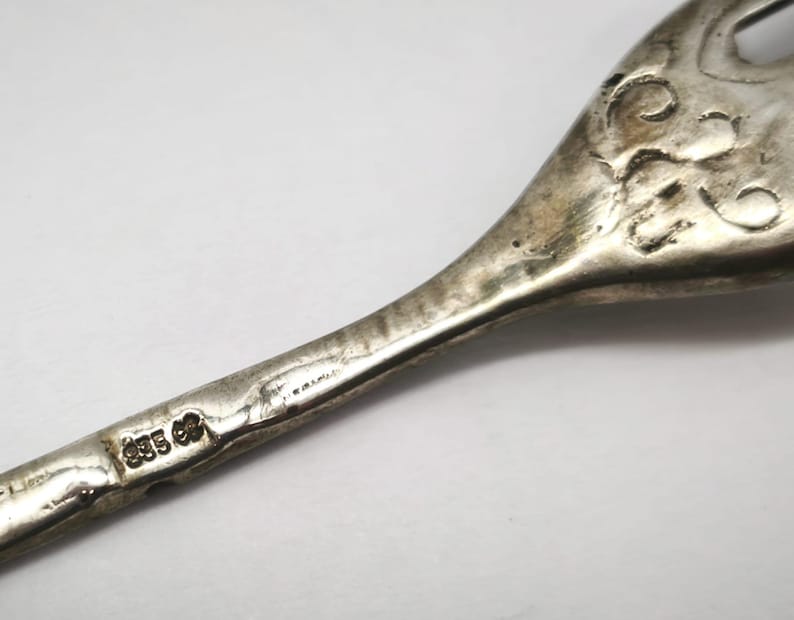 May include: Close-up of a silver serving utensil, possibly a fork or spoon, with ornate scrollwork and a hallmark stamp. The utensil has a tarnished appearance, suggesting age. The hallmark reads "835" and "CP".