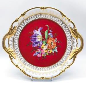 Può includere: Un vassoio da portata decorativo con un vivace design floreale. Il vassoio presenta un centro rosso con un bouquet di fiori colorati, tra cui fiori viola, arancioni e gialli. Il bordo è bianco con dettagli dorati e manici dorati ornati.