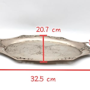 Puede incluir: Una bandeja plateada de forma ovalada con asas decorativas. La bandeja mide 32,5 cm de largo, 20,7 cm de ancho y 1,5 cm de profundidad. La bandeja tiene un dise&ntilde;o ornamentado alrededor de los bordes y probablemente es de metal.
