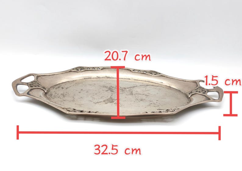 Puede incluir: Una bandeja plateada de forma ovalada con asas decorativas. La bandeja mide 32,5 cm de largo, 20,7 cm de ancho y 1,5 cm de profundidad. La bandeja tiene un dise&ntilde;o ornamentado alrededor de los bordes y probablemente es de metal.