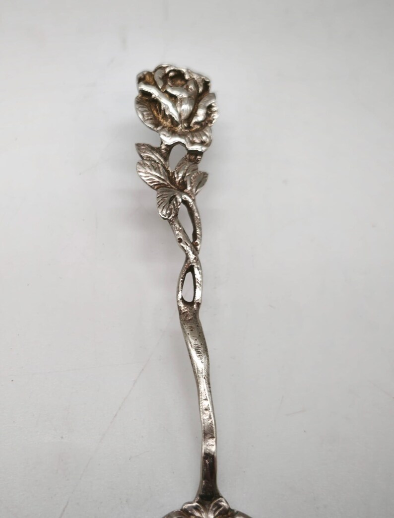 Peut inclure: Une cuill&egrave;re de service argent&eacute;e avec un motif de rose. La cuill&egrave;re pr&eacute;sente une fleur de rose d&eacute;taill&eacute;e en haut, avec des feuilles et une tige incurv&eacute;e. La poign&eacute;e a un design torsad&eacute; d&eacute;coratif.