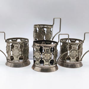 Può includere: Set di tre porta bicchieri da tè in metallo argentato con disegni intricati. Ogni porta bicchiere ha un manico e una base rotonda.