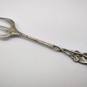 May include: A silver serving fork with three tines. The handle features a floral design with leaves and a flower at the end. The fork is made of a shiny, metallic material and is set against a white background.