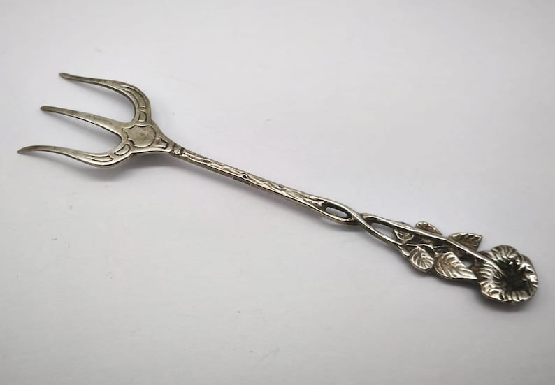 May include: A silver serving fork with three tines. The handle features a floral design with leaves and a flower at the end. The fork is made of a shiny, metallic material and is set against a white background.