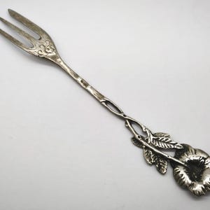 May include: Silver-toned serving fork with three tines and a decorative floral handle. The handle features a flower and leaves design. The fork is likely made of metal and has an antique or vintage appearance.