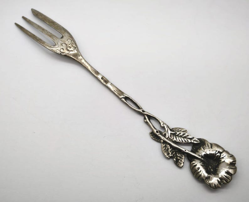 May include: Silver-toned serving fork with three tines and a decorative floral handle. The handle features a flower and leaves design. The fork is likely made of metal and has an antique or vintage appearance.