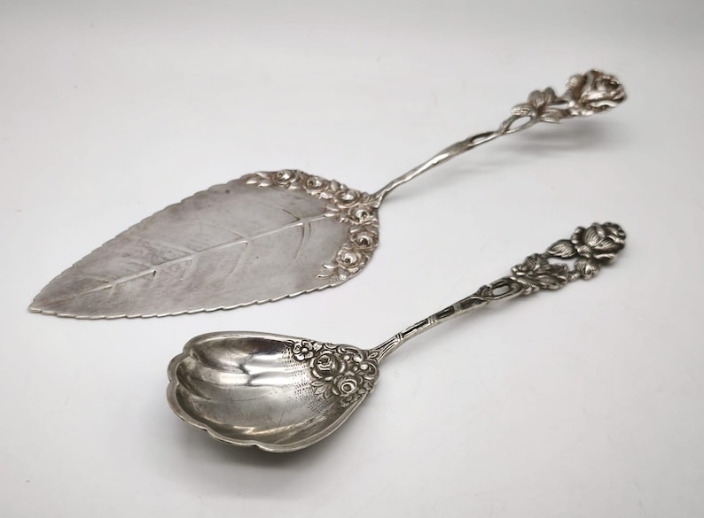Peut inclure: Ensemble de service argent&eacute; comprenant une pelle &agrave; g&acirc;teau en forme de feuille et une cuill&egrave;re. Les deux pi&egrave;ces pr&eacute;sentent des d&eacute;tails floraux orn&eacute;s sur les manches et les bords. La pelle a une grande feuille.