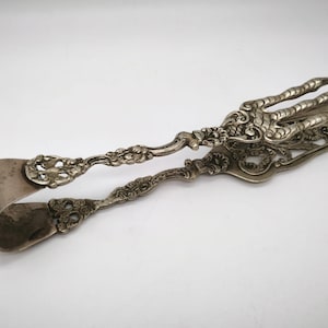 Puede incluir: Pinzas de azúcar plateadas ornamentadas con un diseño floral y de volutas detallado. Las pinzas presentan un extremo de agarre en forma de garra y un mango curvado. El metal tiene un aspecto ligeramente envejecido.