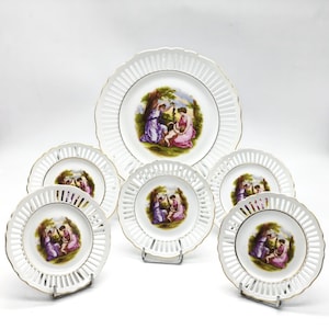 Könnte beinhalten: Sechsteiliges Set weißer Porzellanteller mit goldener Verzierung und einem zarten Blumendesign. Die Teller zeigen ein zentrales Bild von drei Frauen in einer Gartenlandschaft.