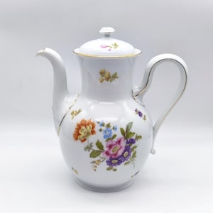 Pode incluir: Cafeteira de porcelana branca com tampa, bico e alça curvos. A cafeteira é adornada com desenhos florais coloridos em laranja, azul, rosa e roxo, com detalhes dourados. Uma peça clássica para servir bebidas.