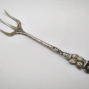 May include: Silver serving fork with three tines and a decorative floral design. The handle features a twisted pattern and a rose motif at the end. The fork is likely made of sterling silver.