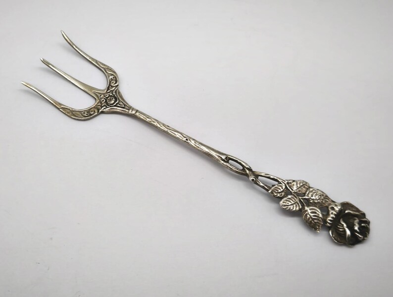 May include: Silver serving fork with three tines and a decorative floral design. The handle features a twisted pattern and a rose motif at the end. The fork is likely made of sterling silver.