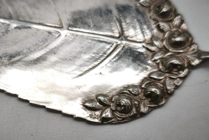 Peut inclure: Gros plan d'une pi&egrave;ce de service en argent en forme de feuille, avec des nervures d&eacute;taill&eacute;es. Le bord est orn&eacute; de roses en relief. La pi&egrave;ce a une surface brillante et r&eacute;fl&eacute;chissante et semble &ecirc;tre antique ou vintage. Le chiffre "800" est estampill&eacute; sur la feuille.