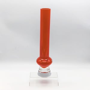 Pode incluir: Um vaso de vidro laranja vibrante, alto, com uma base bulbosa e um caule cilíndrico transparente. O vaso é exibido em um suporte de acrílico transparente, mostrando seu design elegante e moderno. O vaso tem cerca de 30 cm de altura.