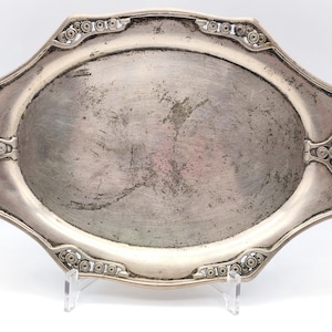 Puede incluir: Bandeja ovalada plateada con asas decorativas y detalles ornamentados. La bandeja tiene un aspecto ligeramente desgastado, con desgaste visible en la superficie. Los bordes presentan un patr&oacute;n circular repetitivo. La bandeja est&aacute; soportada por un soporte transparente.