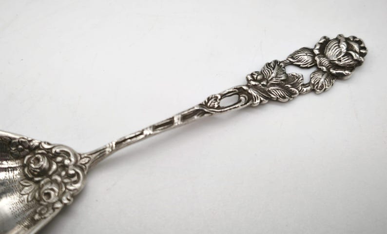 Peut inclure: Une cuill&egrave;re en argent avec un motif de rose sur le manche et le bol. La poign&eacute;e pr&eacute;sente des sculptures d&eacute;taill&eacute;es de roses et de feuilles, ajoutant une touche d&eacute;corative. La cuill&egrave;re est probablement en argent sterling ou en m&eacute;tal similaire.