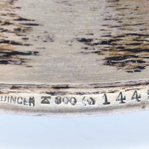 Puede incluir: Primer plano de un objeto de metal plateado con el texto "J.M.HEINRICHDINGER" y los n&uacute;meros "800" y "144568" estampados. El objeto tiene una superficie texturizada.