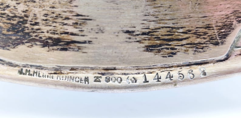 Puede incluir: Primer plano de un objeto de metal plateado con el texto "J.M.HEINRICHDINGER" y los n&uacute;meros "800" y "144568" estampados. El objeto tiene una superficie texturizada.