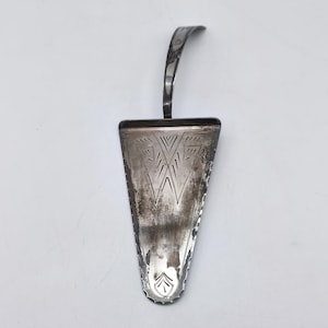 May include: A vintage silver cake server with a triangular blade and a curved handle. The blade features etched geometric designs and a decorative edge. The server is tarnished, showing signs of age and use.