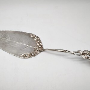 Peut inclure: Une pelle &agrave; g&acirc;teau argent&eacute;e avec une lame en forme de feuille et une poign&eacute;e orn&eacute;e d'une rose. La lame pr&eacute;sente un d&eacute;tail de nervure centrale et est bord&eacute;e de petits motifs floraux. La poign&eacute;e a la forme d'une tige.