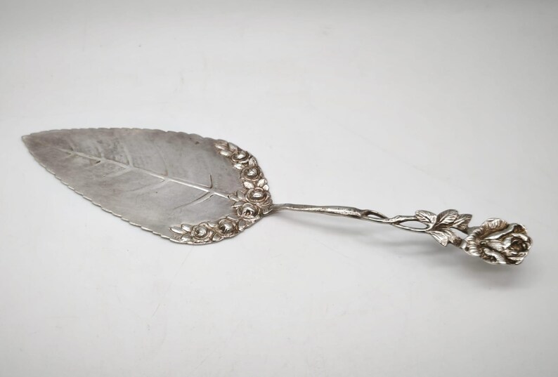 Peut inclure: Une pelle &agrave; g&acirc;teau argent&eacute;e avec une lame en forme de feuille et une poign&eacute;e orn&eacute;e d'une rose. La lame pr&eacute;sente un d&eacute;tail de nervure centrale et est bord&eacute;e de petits motifs floraux. La poign&eacute;e a la forme d'une tige.