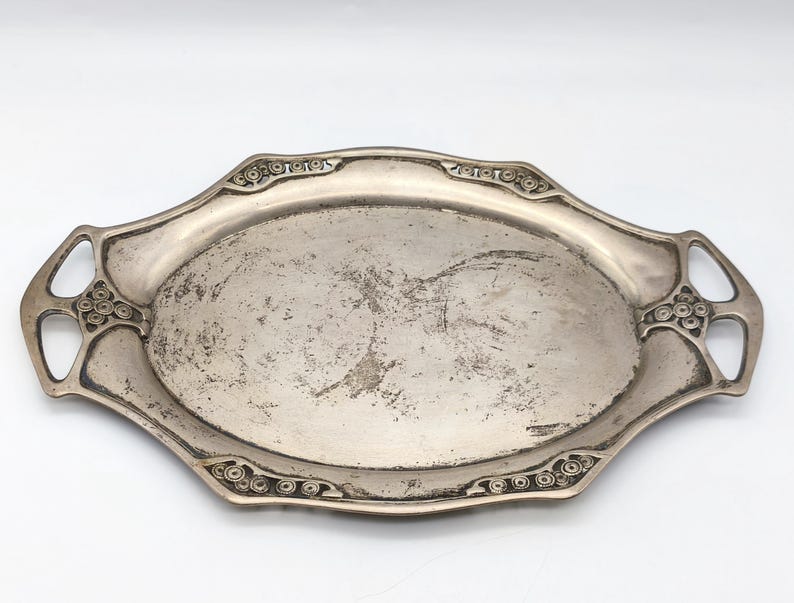 Puede incluir: Una bandeja de servir antigua de color plateado con forma ovalada y asas decorativas. La bandeja presenta detalles ornamentados a lo largo de los bordes y asas, con un acabado ligeramente desgastado. La bandeja est&aacute; vac&iacute;a y lista para usar.