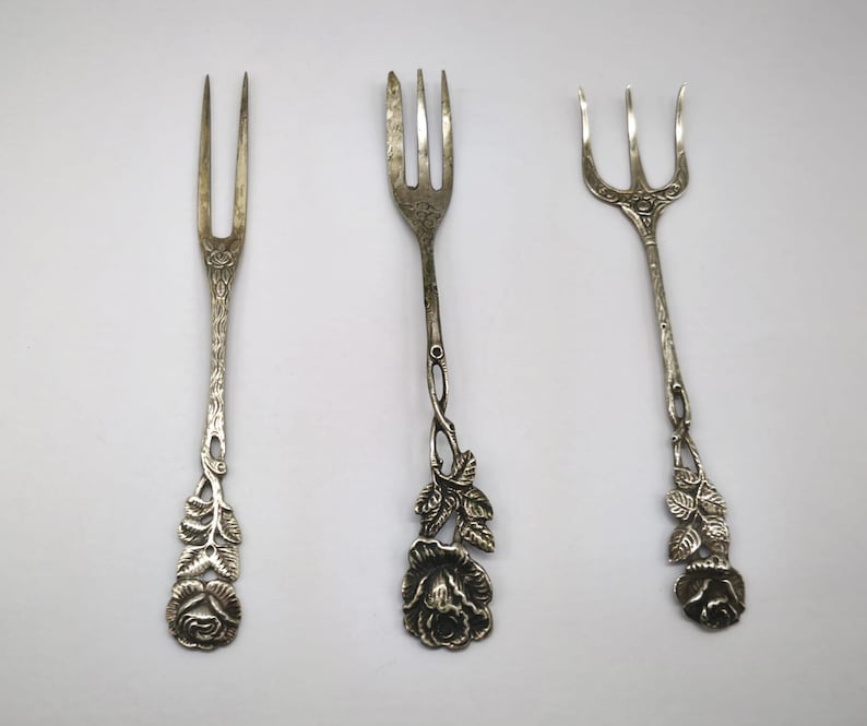 May include: Three antique silver forks with ornate floral designs. Each fork features a different handle design, with rose and leaf details. The forks are displayed on a white background, showcasing their intricate craftsmanship.
