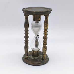 Könnte beinhalten: Eine antike Sanduhr mit einem messingfarbenen Rahmen und klaren Glaskolben. Die Sanduhr wird von vier verzierten Beinen getragen und hat eine kreisförmige Ober- und Unterseite. Der Sand im Inneren ist weiß.