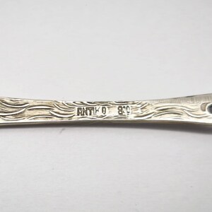 May include: Close-up of a silver antique utensil with intricate leaf and wave patterns. The word "ANTIKO" and the number "800" are stamped on the handle. The utensil has a decorative design.